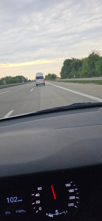 The image captures a view from inside a car on a highway, likely in Sarstedt, Germany, judging by the context provided. The perspective is from the driver's seat, looking forward through the windshield. The car is in motion, as evidenced by the blurred road and the speedometer reading.

In the foreground, the dashboard of the car is visible, with a digital speedometer displaying "102 km/h". Below this, the outdoor temperature is indicated as "21°C" and the odometer reads "91027 km". To the right of the speedometer is an analog dial with a red needle pointing between "160" and "180", suggesting the car's current speed is around 170 km/h, which contradicts the digital reading. There is also a fuel gauge indicating approximately half a tank.

On the highway ahead, a white van is traveling in the same direction, appearing as a distinct object against the overcast sky. The highway has multiple lanes, marked by white lines. To the sides of the highway, trees and foliage are visible, suggesting a rural or semi-rural setting. The sky is cloudy with some lighter patches indicating the direction of the sun. The overall impression is a snapshot of a drive on a German autobahn.