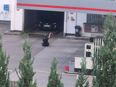 The image depicts an outdoor scene at what appears to be a car wash and gas station. In the background, a black car with the license plate "KL XC 4" is partially visible inside a car wash bay. To the left of the car wash entrance, a blue wall-mounted unit with hoses is present. In the foreground, a person is sitting on the ground facing away from the camera, with their right arm raised. To the right of the car wash area, there are several gas pumps. A red bucket is visible near one of the pumps. The ground surface is paved with interlocking bricks and concrete. Several evergreen trees are scattered throughout the scene. The overall environment suggests a commercial establishment in an urban or suburban setting.