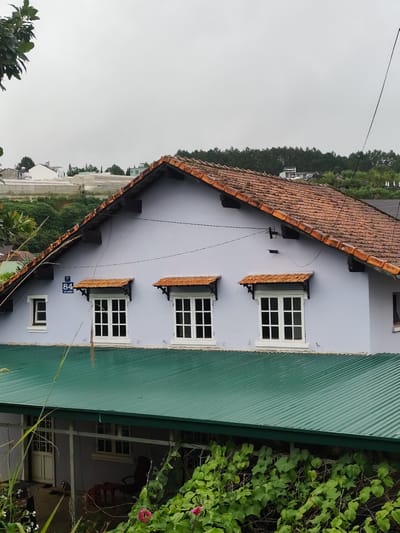 The image depicts a house in Lang Biang - Đà Lạt, Vietnam, under an overcast sky. The main structure has a light blue facade and a red tile roof. Three windows with orange awnings are visible on the upper level, each with a white frame and multiple panes. To the left of these windows, a smaller, single-paned window also has a white frame. A blue street sign with white numbering and text is mounted to the left of the central window. A green corrugated metal roof covers a lower section of the house, extending forward and partially obscuring the ground level. Visible in this lower area are a white-framed door, some furniture including a chair, and dense green foliage with pink flowers in the foreground. The background shows more greenery, including trees and what appear to be greenhouses.