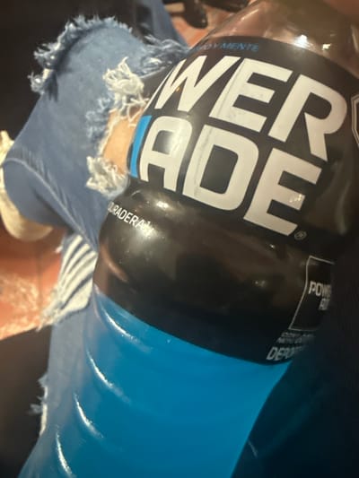 A Powerade bottle with a blue liquid inside rests on a lap covered by distressed denim jeans. The bottle's black label displays the word "POWERADE" in large, white letters. The background is indistinct, but it appears to be indoors. There is no clear evidence to suggest this image was taken in San Cristóbal, Mexico.