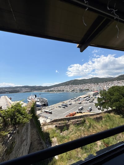 The image captures a scenic view of the port of Kavala, Greece, from a slightly elevated vantage point, likely a balcony or rooftop. In the foreground, a dark, horizontal railing runs across the bottom of the frame, partially obscuring a textured, grey stone wall with patches of green vegetation growing on it. Above this, a dark, corrugated awning with visible support beams extends across the upper portion of the image, with decorative string lights hanging from it.
The main subject of the image is the bustling port area. A large, multi-deck ferry, primarily white with blue accents and marked with "Superfast" branding, is docked at a pier. The ferry has its ramp extended, suggesting it is either loading or unloading vehicles and passengers. Adjacent to the ferry is a large, paved parking lot filled with various vehicles, including buses, vans, and smaller cars. Some orange containers or vehicles are also visible in the lot.
Beyond the port, the city of Kavala spreads out across a hilly landscape. Numerous buildings with red-tiled roofs are densely packed together, climbing up the hillsides. The city is situated along the coast, meeting the clear blue water of the Aegean Sea. In the distance, more hilly terrain, partially covered in greenery, forms a backdrop to the city. The sky is a vibrant blue with scattered white cumulus clouds. The overall impression is one of a lively Mediterranean port city on a bright, sunny day.