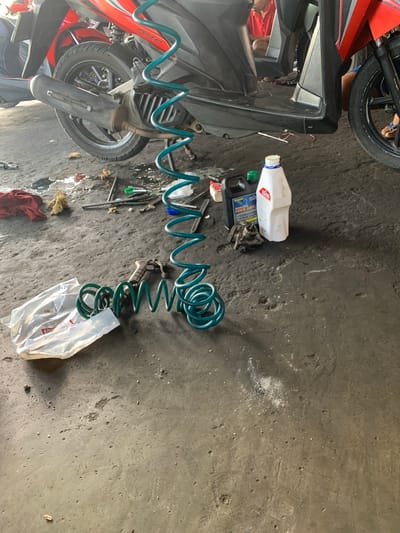 The image depicts a workshop or garage scene in Karangkemiri, Indonesia. A red and black motorcycle is the central subject, partially elevated, revealing its undercarriage. Tools and car maintenance supplies are scattered on the rough concrete floor, which is stained with oil and dirt. Among the items visible are a coiled green air hose, a spray gun, bottles of fluid, and plastic packaging. A person wearing a red shirt is partially visible in the background, suggesting ongoing work or activity within the space. The environment appears to be a utilitarian workspace.