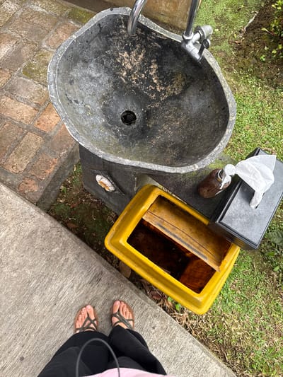 The image shows an outdoor sink with a faucet, a yellow trash bin, a tissue dispenser, and a small brown bottle, all situated on a stone pedestal. The sink is made of rough, dark material and appears weathered. A person's feet, wearing sandals, are visible at the bottom of the frame, standing on a concrete path. The surrounding environment includes grass and brickwork. The scene is located in the city of Banjar, Indonesia, likely in a public outdoor area such as a park or garden.