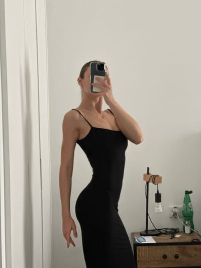This image depicts an indoor mirror selfie taken by a woman in Ericeira, Portugal. The woman is wearing a form-fitting black sleeveless midi-dress with thin straps and a straight neckline. Her face is obscured by a dark-cased smartphone held in her right hand, positioned to capture the reflection. A delicate gold-colored necklace is visible around her neck. The background of the reflection consists of a plain white wall. A white door frame is visible on the left side of the frame. On the right, a dark wooden bedside table or dresser with two black-handled drawers is present. Objects on the table include a black desk lamp with a wooden arm and an exposed light bulb, a tangled black cable, a light green plastic bottle, and minor indeterminate items. A white wall outlet is visible above the table. The woman is posing, slightly turned to her left, for the photograph.