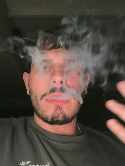 The image depicts an adult male from the chest up, positioned in an indoor setting within the city of Hudson, Argentina. The subject, a man with light skin, brown hair, a beard, and a mustache, is shown exhaling a substantial cloud of white vapor or smoke, which originates from his mouth and obscures parts of his face, including his eyes and forehead. A silver-colored ring is visible in his right nostril. He wears a dark grey long-sleeved shirt with partial white text, "Ligue" and "ancophone de," visible on the chest area. A thin silver chain is visible around his neck. His left hand, blurred due to motion, is partially visible on the right side of the frame, with fingers extended. The background is uniformly dark and featureless, indicating low light conditions.