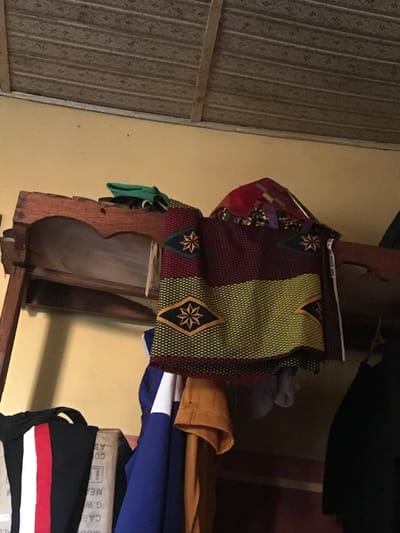 A shelf displays a mix of fabrics and clothing. A colorful, patterned fabric with star-like designs is stacked on top. Below, a glimpse of mustard-colored fabric peeks out from beneath a folded blue and white garment. The shelf's wooden structure has a unique curved design. A glimpse of black fabric with a white and red stripe sits at the bottom. To the left, a patterned metal door or wall is visible.