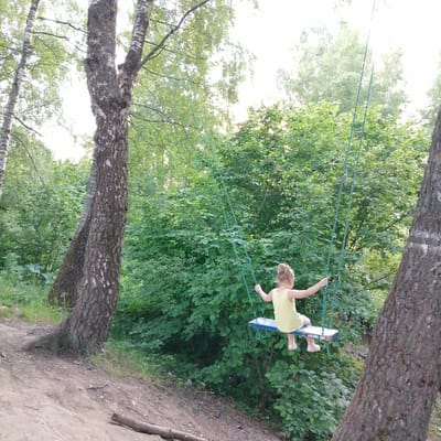 A young girl with blonde hair pulled into a ponytail, wearing a yellow sleeveless top and white shoes, is seated on a blue and white swing. She is holding onto the green ropes of the swing, her body leaning slightly forward. The swing is suspended between two trees, with the girl positioned in front of a dense wall of green foliage. The background consists of more trees and vegetation, with dappled sunlight filtering through the leaves. In the foreground, a dirt path slopes upwards, with a fallen log visible on the path. Two large tree trunks with textured bark frame the scene on the left and right sides. The overall environment appears to be a natural setting, likely a park or wooded area. The image was captured in Kryukovo, Russia.
