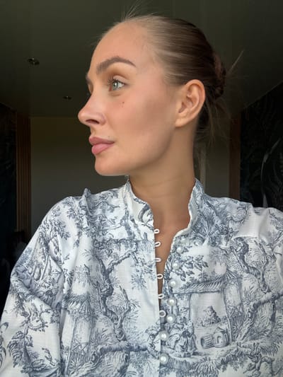 The image is a close-up portrait of a young woman with light skin and light brown hair pulled back in a bun. She is shown in profile, facing left, with her head tilted slightly upwards. Her eyes are light blue-green, and she has light makeup, including blush and lip color. There are a few small dark spots on her cheek. She is wearing a white shirt with a blue and white toile print. The shirt has a high, ruffled collar and is fastened with pearl buttons. The background is dark and appears to be an indoor setting with a window to the left, offering a glimpse of what might be wooden paneling or blinds. The overall lighting suggests it might be daytime, possibly near a window. The scene is devoid of any specific elements that would identify it as being in the city of Шолохово, Russia.
