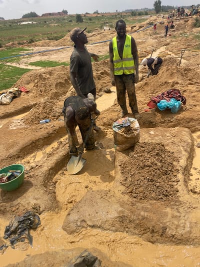 The image depicts a muddy, disturbed area in Maiyanga, Nigeria, with three individuals present. One man wears a high-visibility vest with "SAIO" printed on it. Another man is bent over, shoveling mud, while the third individual stands nearby. Mud and water cover much of the ground. Personal items such as clothing, bags, and a water bottle are scattered around the site. The background shows a landscape with visible signs of activity.
