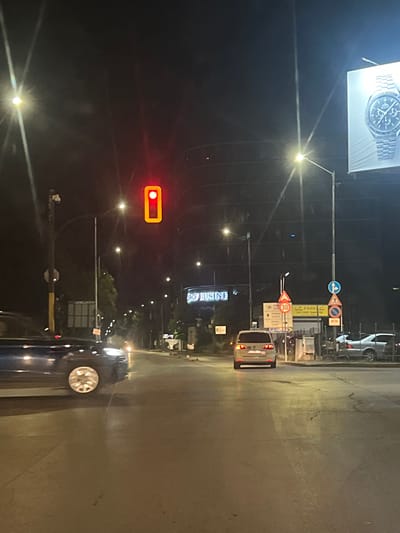 The image depicts a street scene at night in Krivina, Bulgaria. A traffic light with a red signal is visible in the foreground. A dark-colored vehicle is in motion, partially obscuring the road. Further down the street, a silver minivan is also in motion, its brake lights illuminated. To the right, a large billboard displays a watch advertisement. Several streetlights illuminate the area, and various road signs are present, including a speed limit sign indicating 30. The environment appears to be an urban or suburban setting with buildings and street infrastructure visible.