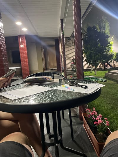 The image shows a close-up view of a glass-topped table outdoors at night. On the table are several papers, a set of keys with a rainbow-colored keychain, and a colorful striped object, possibly a wallet or small bag. The table is made of black metal and has a textured glass surface. In the background, a grassy lawn is visible, along with some pink flowers in a planter. A building with red brick accents and outdoor lighting can also be seen. The scene appears to be a backyard or patio area. The overall impression is that of a casual evening gathering or activity taking place in a residential setting. The time of day is clearly night, indicated by the artificial lighting and the darkness of the surroundings. The weather appears calm, with no indication of rain or wind. There is no visible text in the image.