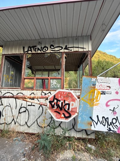 The image depicts an abandoned, graffiti-covered structure in Perazića Do, Montenegro, under a clear blue sky. The structure, possibly a former toll booth or guard house, has a corrugated metal roof and siding. Its windows are broken and boarded with wire mesh. Numerous spray-painted tags and graffiti adorn the walls, a rusted metal gate, and various signs attached to the structure. These signs include a red octagonal stop sign with black graffiti, a blue metal sign with handwritten text and yellow spray paint, and a white board with multiple graffiti tags and stickers, one of which reads "FARGO TIME". Overgrown vegetation and debris are present at the base of the structure, indicating its derelict state. The background features a hillside covered with trees, suggesting a semi-rural or natural setting within the town.