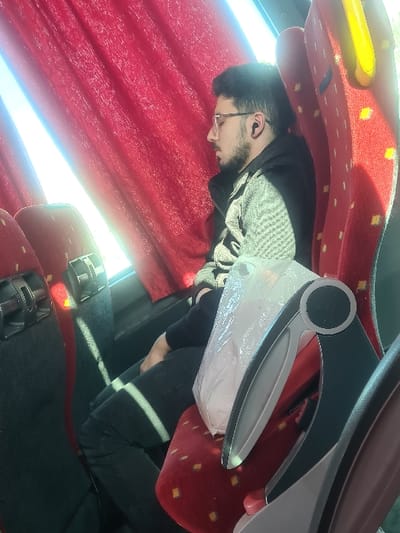 Inside a passenger vehicle, presumed to be a bus, in Al-Kiswa, Syria, a single male subject is depicted. The subject, appearing to be an adult male with dark hair and a beard, is seated on a red upholstered passenger seat with gold diamond-patterned accents, positioned next to a window. Red fabric curtains cover the window. He is wearing eyeglasses and has a black earbud in his right ear. His eyes are closed, indicating he is sleeping or resting. He is dressed in a dark jacket over a grey patterned sweater and dark trousers. His right arm is visible, resting on his lap. Directly in front of him, a white translucent plastic bag is resting on the backrest of the forward seat, which also features a grey and black plastic tray or handle mechanism. Bright sunlight illuminates the scene from the window side, casting distinct diagonal light patterns across the interior, including the floor and the subject's leg. Multiple rows of similar red passenger seats are visible within the vehicle's interior.