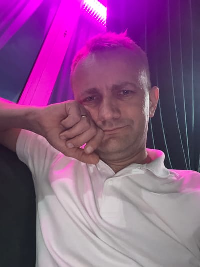 This media file shows a close-up, horizontally oriented portrait of a man. He is wearing a white polo shirt and has his right hand covering his mouth. A ring is visible on his left hand. The man appears to be looking directly at the camera with a slight smile. The background is dark and features horizontal lines, possibly indicating a textured surface or seating. To the left of the man, a bright pink light is illuminating the scene, creating a vibrant glow on his face and shirt. The lighting suggests an indoor setting, possibly at night or in a dimly lit environment. No text is visible in the image. The overall mood is relaxed and intimate.