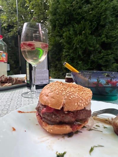 A close-up shot captures a gourmet burger and a glass of white wine on a table set for an outdoor meal. The burger, served on a white plate, is topped with sesame seeds and appears to be made with a thick, juicy patty and possibly some red sauce and pickles. To the left of the burger, a wine glass filled with white wine and garnished with berries sits next to a bottle of alcohol, likely vodka, given its dark color and label which suggests it's from Russia. In the background, a blue bowl filled with salad and a yellow serving utensil can be seen, along with a green, lush hedge. The setting appears to be a backyard or patio in Pribrezhnyi, Russia, suggested by the presence of Russian alcohol and the general ambiance of an outdoor gathering.