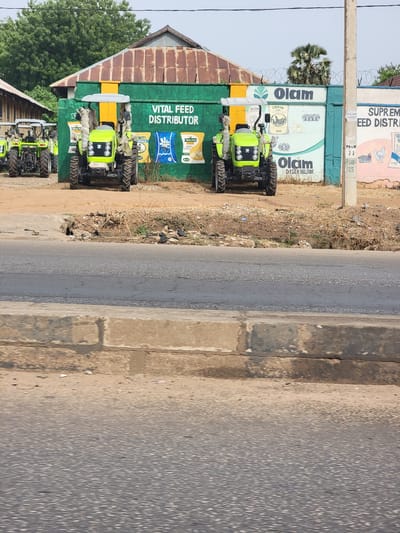 The image depicts a row of green tractors parked in front of a building with advertisements in what appears to be Barnawa, Nigeria. The building is painted with "VITAL FEED DISTRIBUTOR" in white letters and features images of animal feed products. The name "Olam" is prominently displayed on the building as well. The tractors are new and appear to be for sale or distribution. A paved road runs along the edge of the scene. Additional signage for "SUPREME FEED DISTRI" is partially visible. There are no people visible in the immediate scene. The environment appears to be a commercial area.