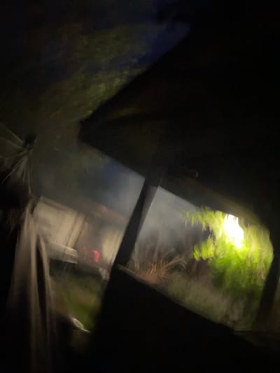 The image captures a dimly lit scene, possibly at night, with elements of an outdoor setting. A large, dark, angled surface dominates the foreground, suggesting a roof or overhang. To the left, a blurred, pale fabric or material hangs down, partially obscuring the view. Beyond this, the scene opens up to a blurry background where faint red lights are visible, indicating a possible fire or artificial light source. Green foliage, illuminated by a bright, diffuse light, is present to the right, creating a contrasting element against the darker surroundings. The overall impression is one of low light conditions, with significant motion blur, making precise identification of objects and their interactions difficult. The location is identified as Драчкаўскі сельскі Савет, Belarus.