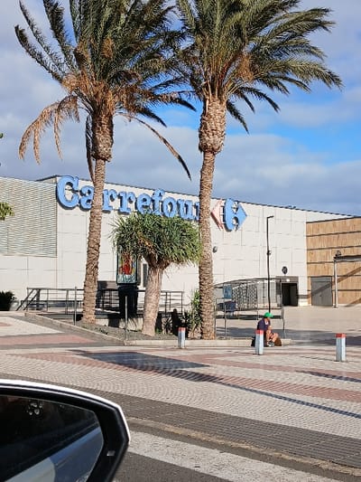 The photo captures a Carrefour supermarket with its blue signage flanked by palm trees under a blue sky. A pedestrian walks on a brick-patterned sidewalk in front of the building. Part of a car's side mirror is visible in the foreground. The perspective is from a low angle, possibly taken from inside a vehicle.