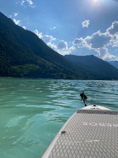 The image depicts a serene lake scene in Lärchenwiese, Austria, as viewed from a paddleboard. The perspective is from the front of the paddleboard, which occupies the right side of the frame. The paddleboard is light gray with a textured diamond grip pattern and has some visible white lettering. A dark rope is attached to the board and disappears into the water.

In the water, a small, dark object, possibly a duck or a water bird, is visible in the distance, breaking the surface of the clear, turquoise water. The water is calm with gentle ripples reflecting the sunlight.

The left side of the image features a majestic mountain covered in dense green forest, with a clear blue sky above. Several fluffy white clouds are scattered across the sky, with the sun peeking through from the upper left, casting a bright glare and creating sunbeams. The mountain slopes down towards the lake, and in the distance, a small settlement with buildings is visible nestled along the lakeshore. The overall scene suggests a sunny day, likely late morning or early afternoon, with pleasant weather ideal for outdoor activities on the water. The atmosphere is peaceful and tranquil.