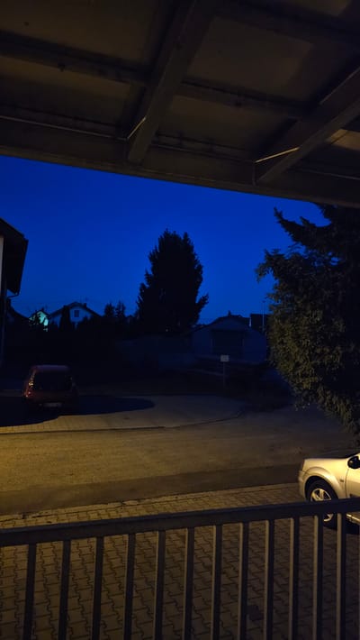 The image depicts a twilight scene in Vöhringen, Germany. The sky is a deep blue, indicating the time is shortly after sunset. In the foreground, a metal railing with vertical bars runs across the bottom of the frame. Behind the railing, a paved area with interlocking bricks is visible. To the left of the paved area, a portion of a road or driveway is present, with a parked red car partially visible. The car has a license plate with the inscription "NU IV B65".
In the middle ground, a residential street curves gently. Houses with illuminated windows are silhouetted against the dark sky. A tall, dark tree stands prominently in the center. To the right, a large, dark evergreen tree is partially visible. Beyond the trees and houses, a low-rise building with a grey facade and a large door is situated.
The overall impression is one of a quiet suburban evening. The lighting suggests artificial illumination from streetlights or house lights, casting long shadows on the ground. The scene is static, with no people or animals visible.