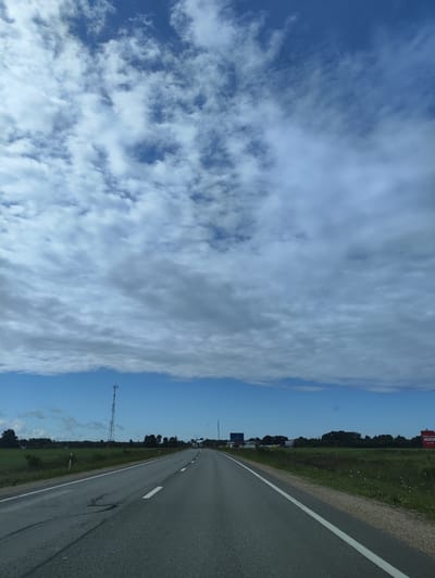 The image shows a view from a car driving on a paved road in Ainaži, Latvia. The road has two lanes and white markings. On the left side of the road, there is a green field and a tall communication tower. On the right side, there is a grassy shoulder and a field with some white flowers. In the distance, there are trees, houses, and road signs. The sky is blue with scattered white clouds. There are no people or animals visible in the image.