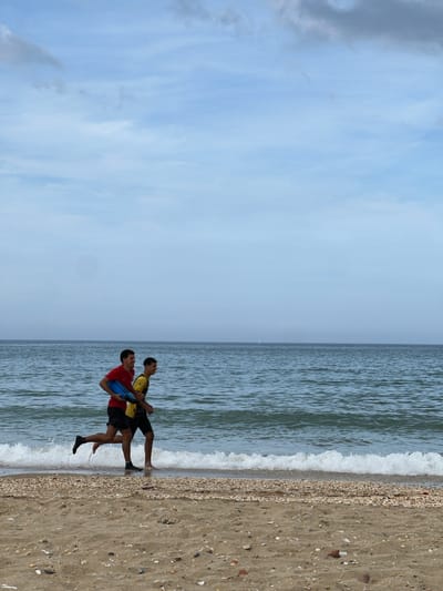 Two individuals are standing at the edge of the water on a beach. One is wearing a red shirt and carrying a blue object, the other is wearing a yellow shirt. The location is M'Diq, Morocco, as evidenced by the beach setting and coastal characteristics. The sky is overcast. Waves are visible at the shoreline. The beach consists of sand and small shells.