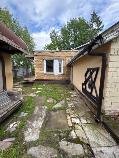 The image shows a slightly overgrown backyard in Naro-Fominsk, Russia. A wooden bench is visible in the foreground, next to a small, stone-clad structure. A pathway made of concrete slabs and some broken tiles leads towards a yellow-colored building with a corrugated metal roof. To the left, a brick building with a light brown facade and a sloping, rusty metal roof is partially visible. Another brick building with yellow bricks and a white window is situated behind the pathway. A dark brown downspout runs along the side of the yellow building. The area is surrounded by trees and a fence in the background. The sky is cloudy with patches of blue.