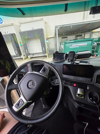 This media file is a photograph taken from inside the cab of a truck, looking out through the windshield and side windows. The interior of the truck is visible, featuring a steering wheel with a Mercedes-Benz emblem, dashboard controls, a digital display screen, and a smartphone mount. A charging cable is plugged into the dashboard.

Outside the truck, the scene appears to be an industrial area or loading dock. There are large, white loading bays visible, some marked with numbers "13" and "14". A green truck or waste disposal vehicle with the text "KNETTENBRECH GURDULIC" and "Husmann" is parked next to the truck. Traffic lights, showing green and red signals, are also visible near the loading bays. The overall environment suggests a daytime setting, possibly a logistics hub or a facility where goods are being loaded or unloaded. The image is rotated 90 degrees counter-clockwise.