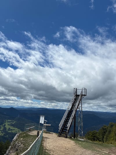 The image depicts a tall metal structure resembling an observation tower on the edge of a cliff or mountain. The tower consists of a staircase leading to a platform at the top. In the background, there's a landscape of mountains and sky with clouds. A portion of the landscape is on the right side of the image, consisting of trees and dirt. A sign is partially visible on the right side of the image. The location is Giovren, Bulgaria.