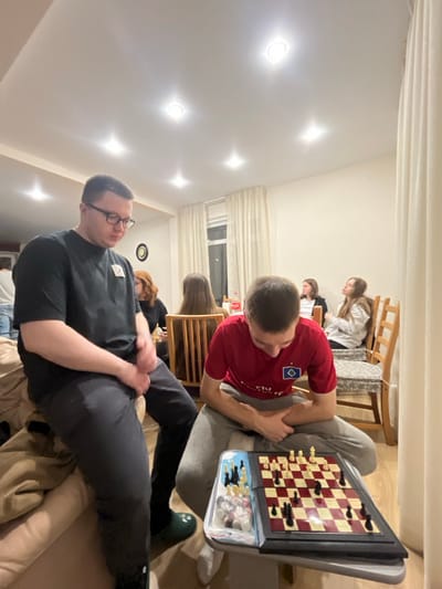 The image displays an indoor scene located in Заозерное Снт, Russia, depicting a social gathering. In the foreground, two males are prominent. The male on the left, wearing spectacles, a black short-sleeved t-shirt, and dark trousers, is seated on the edge of a light-colored sofa, looking downwards with his hands clasped. He wears a dark green slip-on shoe on his visible right foot. The male on the right, clad in a red short-sleeved t-shirt featuring a white Hamburg SV logo and light grey sweatpants, is seated and intently focused on a chess board placed on a small table before him. The chess board has red and off-white squares with both black and white pieces actively set for play; additional chess pieces are contained in a clear plastic bag next to the board.
In the mid-ground and background, approximately five other individuals are visible, primarily seated at a wooden dining table with chairs. A woman with reddish-brown hair is partially visible to the left of the table. A red Coca-Cola bottle rests on the table. The room features light-colored walls, a wooden floor, and a white ceiling equipped with multiple recessed downlights. A large window with white curtains is visible in the background, showing a dark outdoor environment. A round, dark object is mounted on the far wall.