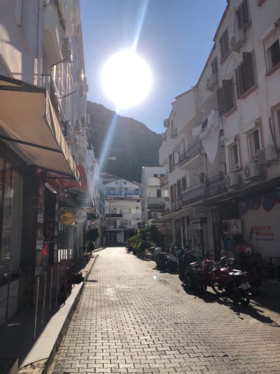 The image shows a narrow street in Kaş, Turkey, lined with multi-story buildings. The street is paved with interlocking bricks and is illuminated by bright sunlight, creating strong shadows. On the left side of the street, there are awnings extending from shops, with some chairs and tables visible. On the right side, buildings feature balconies with air conditioning units and some laundry hanging. Several motorcycles and scooters are parked along the street. The street recedes into the distance, leading towards a hillside. The overall impression is of a sunny day in a Mediterranean town.