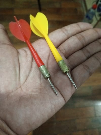 The image presents a close-up, top-down view of two darts held in an open human hand. The hand, with medium skin tone, is positioned with the palm facing upwards, supporting the darts. One dart has a distinct red plastic flight and a red shaft. The second dart features a yellow plastic flight and a yellow shaft. Both darts share identical construction details: a textured, knurled brass barrel, which transitions into a sharp, conical, silver-colored metal point. The background is out of focus, displaying blurred brown and dark tones indicative of an indoor setting, possibly a wooden floor or table. The scene is located in the city of Al Haouafate, Morocco.