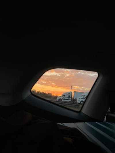 The photo captures a sunset view through a car window. The sky is a mix of orange, yellow, and white clouds reflecting a vibrant sunset. Two white trucks are parked on the roadside, partially obstructing the horizon. The surrounding car interior is dimly lit, almost black, creating a contrasting frame for the bright sunset scene.
