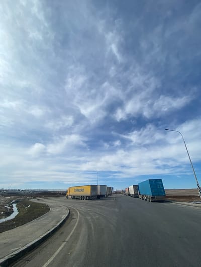 The image shows a scene in Zarya, Russia. The sky is predominantly blue with wispy white clouds. On the right, there is a wide asphalt road curving sharply to the right. Along the road, a line of semi-trailer trucks is parked. A bright yellow truck with the name "OTRASHO" written on its side is clearly visible. Behind it, blue shipping containers are stacked on trailers. A lamppost rises from the side of the road. To the bottom right, a patch of green vegetation with some puddles of water can be seen. The overall environment suggests an industrial or transportation area.