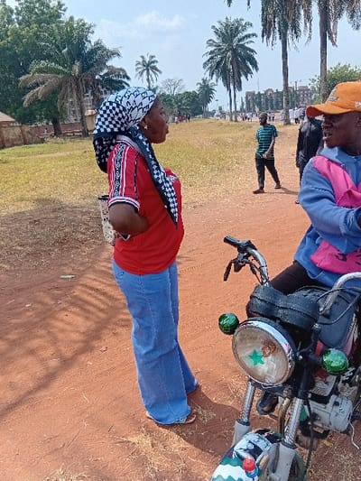 A woman in a checkered headscarf, red shirt, and blue jeans stands in a grassy field with palm trees. Nearby, a man in an orange hat and light blue shirt sits on a motorbike with a green star sticker on the headlight. Another person in a striped shirt walks in the background. The ground is a mixture of grass and reddish-brown dirt.