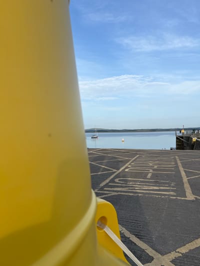 The image depicts a bright, clear day with a vast blue sky streaked with wispy white clouds. In the foreground, a bright yellow object, possibly a bollard or a piece of safety equipment, dominates the lower portion of the frame, curving out of view.

Beyond the yellow object, a body of water stretches towards the horizon, appearing calm and serene. A single sailboat with a white hull and mast is visible on the water. In the distance, a faint shoreline is discernible, suggesting a coastal or lakeside setting. Part of a pier or dock structure can be seen on the far right, with what appears to be a yellow navigational buoy in the middle distance.

The ground in the mid-right of the image is paved with dark asphalt and marked with white lines, forming a diamond pattern and a large, square shape containing the word "PARKING". The lettering is in large, capital white block letters.

The lighting suggests it is daytime, likely late morning or early afternoon, given the brightness and the clarity of the sky. The weather appears to be fair. The location is identified as Terhillion, Ireland. No people are visible in the image. The overall mood is one of peace and openness, with the vast sky and water creating a sense of tranquility.
