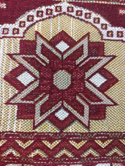 This is a close-up, overhead shot of a woven textile featuring a central eight-pointed star pattern. The star is rendered in a deep red color against a background of yellow and white threads, creating a textured, pixelated effect. The star's points are sharp and angular, with diamond shapes inserted within them, also in red, white, and a pale blue-gray. The center of the star is a small, textured circle in a lighter shade of red.
Surrounding the star is a semicircular border of repeating square motifs, alternating between red and white, set against the yellow and white background. Above this border, another decorative band of red and white squares is visible. Below the central star, a zig-zag pattern in red and yellow, interspersed with green and white threads, forms another decorative border.
The textile appears to be a rug or a similar decorative fabric, likely a prayer rug given the cultural context of Ashgabat, Turkmenistan, which is known for its intricate carpet designs. The lighting is even, suggesting indoor lighting or an overcast day, with no strong shadows. The image focuses solely on the intricate design of the textile, with no people or other objects present. The focus is sharp on the woven threads, highlighting the texture and color variations.