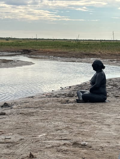 A mud-covered person sits cross-legged on the muddy shore of a body of water. The person is facing to the right, with their head slightly bowed and their arms resting on their lap. The skin of the person is entirely covered in dark mud, obscuring any distinguishing features except for their silhouette and the shape of their body. Their hair appears to be pulled back, also covered in mud, with what look like dark goggles or eye coverings visible. The water in front of them is calm with small ripples. The surrounding environment is a barren, muddy landscape with sparse vegetation in the distance. The ground is cracked and dry in places. In the far background, a flat, green plain is visible under a cloudy sky with thin, horizontal clouds. There are a few distant, thin poles or structures dotted across the landscape. The location is identified as a natural mud pool in the Yamyshevskoye rural district, Kazakhstan.