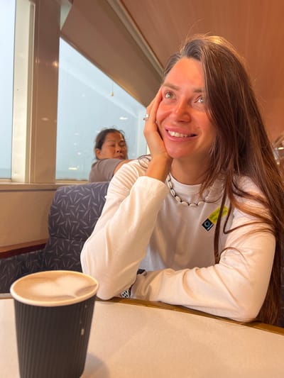 The image shows two people in a brightly lit, indoor setting, possibly a cafe or restaurant with a large window. A woman with long brown hair is in the foreground, leaning on her hand and smiling at the camera. She is wearing a white long-sleeved shirt with a shell necklace and a ring on her finger. A yellow tag with text is visible on her shirt. Behind her, reflected in the window, is another person lying down with their eyes closed, also appearing to be in a casual or resting position. The reflection also shows a patterned fabric on what looks like a seat or cushion. A black cup with a light-colored beverage is placed on the table in the foreground, partially obscuring the woman's arm. The overall mood appears relaxed and casual. The presence of a window suggests it might be daytime. Visible text on the yellow tag appears to be "Samui View" and a phone number. The location context is Don Sak, Thailand.