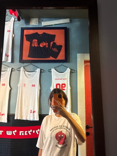 An indoor mirror selfie captures a female individual holding an orange-cased smartphone to obscure her face, while wearing a white t-shirt with a red "BANCO DE SANGRE" graphic. The reflection, framed by a dark mirror, reveals a retail merchandise display against a two-toned wall (blue-gray above, black below), characteristic of a restaurant or themed store environment, identified as "Marvena Burger" located in the Galil Elyon Regional Council, Israel.
The upper blue-gray wall features a large framed display showcasing a black t-shirt with orange graphics of a hamburger and a tractor, alongside a smaller black t-shirt with partial text "SON COW DE". A white rectangular light fixture with a green indicator is mounted above the frame. Below this, three white tank tops are suspended on black hangers, displaying red branding including "oe", "30132", and Hebrew text "מר ונה" (Mar VeNeh). The lower black wall section includes a horizontal red stripe and visible red Hebrew text "מר ונה". To the right of the mirror's reflection, a vertical reddish-orange wall segment with a dark door handle is partially visible.