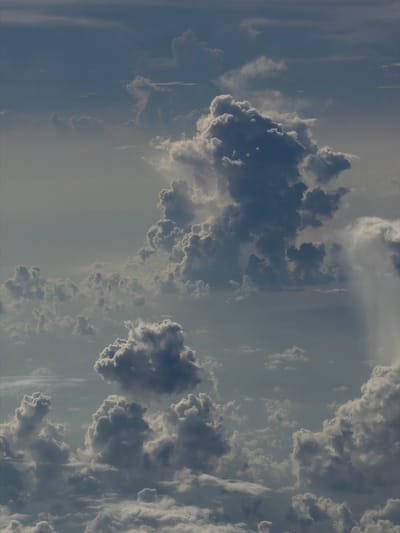 The image depicts a sky filled with cumulus clouds. The clouds are varying shades of gray and white, indicating different densities and levels of sunlight penetration. The background sky is a pale blue. There is no recognizable information to infer the location is Taoyuan City, Taiwan, or to identify specific people, objects, actions, or interactions. The environment is atmospheric.