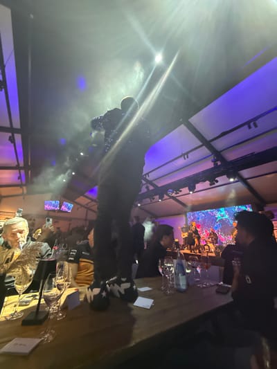 The image captures an indoor event held at night or under low-light conditions within a large tent or temporary structure in Hockenheim, Germany. A person is positioned centrally in the foreground, standing on a wooden table, facing away from the camera towards a distant stage. This individual wears dark, possibly sequined or reflective, clothing on the upper body and dark trousers, paired with distinct black and white chunky sneakers featuring star patterns.

The wooden table in the foreground displays several wine glasses, some containing red liquid, a bottle of sparkling water, and multiple small white cards. To the left of the central figure, an older man with grey hair and a beard is seated at the table, wearing a patterned jacket and exhaling smoke from what appears to be a cigarette or cigar. Behind him, several other individuals are visible, some holding up smartphones. To the right of the central figure, two individuals are seated at the same table, observing the scene towards the stage.

In the background, a live band performs on an elevated stage, featuring at least one vocalist, various musicians, and instruments, including drums and guitars. A large screen behind the band displays colorful, abstract visual graphics. The venue's ceiling, indicative of a large tent or marquee, is illuminated by purple and blue ambient lighting, while bright white stage lights cut through visible atmospheric smoke or haze, creating beams of light.