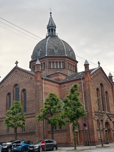 The image shows the facade of a large brick church in Frederiksberg, Denmark, under a cloudy sky. The church features a prominent domed roof with a steeple topped by a cross. Several tall, arched windows are visible on the brick walls. In front of the church, several cars are parked along the street, and young trees with green foliage are planted in rectangular planters. A traffic sign with a red circle and a diagonal line, indicating a prohibition, is visible on the right side of the image. The architecture suggests a historic religious building.