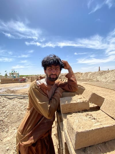 The image shows a man in Deh-E Bagh, Afghanistan. He is standing outdoors in a sandy, dry area, leaning against a stack of unbaked bricks. The man is wearing a long, brown traditional robe with embroidered details along the sleeves and chest. He has dark hair and a beard and is looking directly at the camera, with his right hand resting on his head. In the background, there are stacks of bricks, a small green structure with flags, a red motorbike, and open fields. The sky is blue with scattered white clouds. The environment suggests a brick-making yard or an area where bricks are being produced.