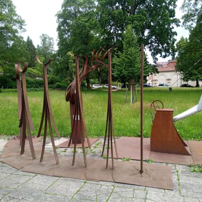 The image shows a public art installation in the city of Eisenach, Germany, likely in a park or open space. The installation consists of several abstract sculptures made of weathered steel. In the foreground, there are at least five tall, slender metal figures, some with curved elements at the top resembling arches or horns. These figures are set on square metal bases, which in turn rest on a paved walkway made of grey stones. Behind the sculptures, a large grassy area is visible, dotted with small yellow flowers and patches of greenery. In the middle ground, trees with lush green foliage form a backdrop, and beyond them, several cars are parked on a road. Further in the distance, a building with a red tiled roof and arched windows is partially visible, suggesting an urban setting or a building adjacent to the park. The sky is overcast.