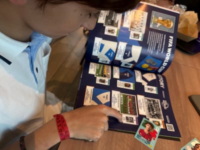 A person is seen indoors, likely in Matosinhos, Portugal, engrossed in a FIFA World Cup sticker album. Their right hand points to a section within the album, while an unpeeled Panini sticker featuring Portuguese footballer João Félix rests on the open page. The individual wears a white polo shirt with blue accents and a red fabric wristband that has faded text, including "DOSE...".
The open sticker album displays pages dedicated to the "FIFA WORLD CUP STORY," featuring sections with text such as "SHARING THE MAGIC OF FOOTBALL" and an explanation of the FIFA Museum's role in preserving football heritage. Various panels are labeled "FWC 6," "FWC 10," "FWC 12," and "FWC 13," corresponding to historical World Cups, including "1966 WORLD CUP ENGLAND," "1970 WORLD CUP BRAZIL," "1974 WORLD CUP GERMANY FR," and "1978 WORLD CUP ARGENTINA." These sections contain small images of teams and players, with some designated "TOP SCORER." A prominent image of the FIFA World Cup trophy and a "FIFA MUSEUM" logo are also visible on the page. A QR code is present in the top left of the album. The activity suggests collecting or reviewing football memorabilia in a casual setting with a wooden surface, possibly a table or floor.
