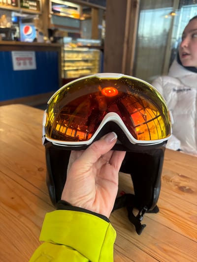 The scene is set indoors at an establishment in Vasil'evo, Russia, resembling a ski lodge cafe or cafeteria. In the immediate foreground, a person's left hand, with manicured fingernails, holds a black ski helmet adorned with white-framed goggles. The goggles feature highly reflective, amber-tinted lenses, prominently displaying a circular red reflection near the top center, likely from an overhead light fixture. The lenses also reflect vertical window panes and portions of the interior space. The person's left forearm, clad in a bright neon yellow puffy sleeve, is visible below the hand.
The objects rest on a light-toned wooden table. In the midground, to the right, a young female individual wearing a white padded jacket is partially visible from the chest up, looking towards the right, outside the frame. Her mouth is slightly open. The background on the left features a blue counter, behind which a silver Pepsi soda dispenser is visible. Above the counter are shelves stocked with various bottles. To the right of the counter, a glass display case with multiple shelves holds numerous pastries or baked goods. Large windows or glass panels extend across the right side of the background, reflecting interior light sources.