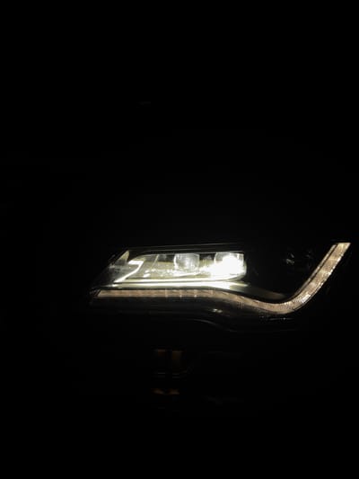A close-up, low-light photograph shows a car's LED headlight. The headlight is illuminated, emitting a bright white light that highlights the intricate design of the light assembly. The surrounding car body is dark, creating a stark contrast that draws attention to the headlight. The background is completely black, suggesting a nighttime or indoor setting. The car's location in Jablonné nad Orlicí, Czechia, is not discernible from the image itself.