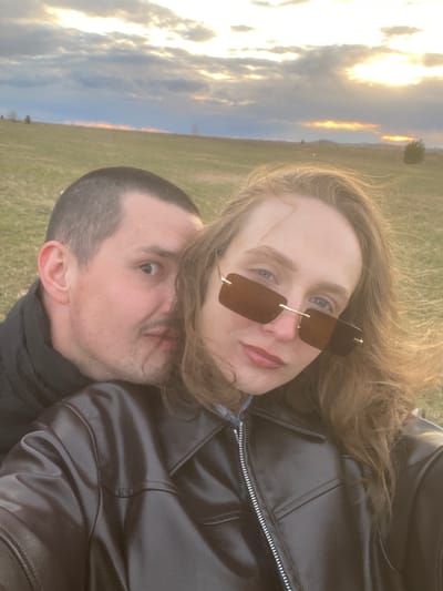 The image features two individuals, a woman and a man, in a close-up outdoor portrait. The photo appears to have been taken during golden hour (sunrise or sunset) in Kueda, Russia, set against a backdrop of a grassy field and a dramatically lit sky.
The woman is positioned in the foreground, slightly above and in front of the man. She has long, wavy light brown hair and is wearing rectangular brown-tinted sunglasses with a delicate gold-colored frame, partially obscuring her blue eyes. Her lips are painted a reddish-pink, and she looks directly towards the viewer with a neutral to slightly smiling expression. She is dressed in a black jacket, possibly leather, which has a visible silver zipper.
The man is situated behind and slightly below the woman. He has short, dark hair and stubble, with his ear prominent. He looks upwards and to his right (from his perspective) with an expression that appears somewhat surprised or curious. He is also wearing a black jacket.
The background shows an expansive sky with scattered clouds, intensely illuminated by the low sun. This creates warm golden and orange hues, particularly along the horizon line. A faint, distant tree can be seen against the brightly lit sky. The setting appears to be an open, grassy field. The overall impression is a casual and intimate shared moment outdoors. No text is visible in the image.