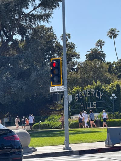 The image shows a daytime view in Beverly Hills, United States of America. A traffic light displays a red signal. A street sign indicates the location as N. Santa Monica Blvd 9400C. In the background, the iconic "Beverly Hills" sign is visible with a group of people standing near it. Some people are taking photos of the sign. Trees and grass surround the area. A black SUV is parked in the foreground.